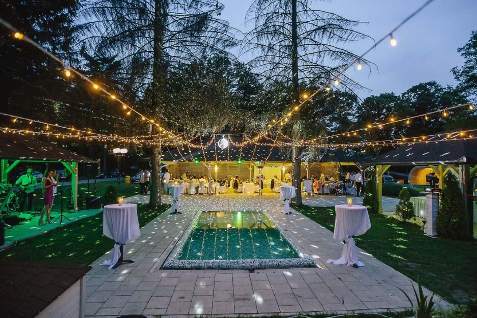 Evening party setup with a pool, string lights and high tables at a bachelor party venue in Manesar